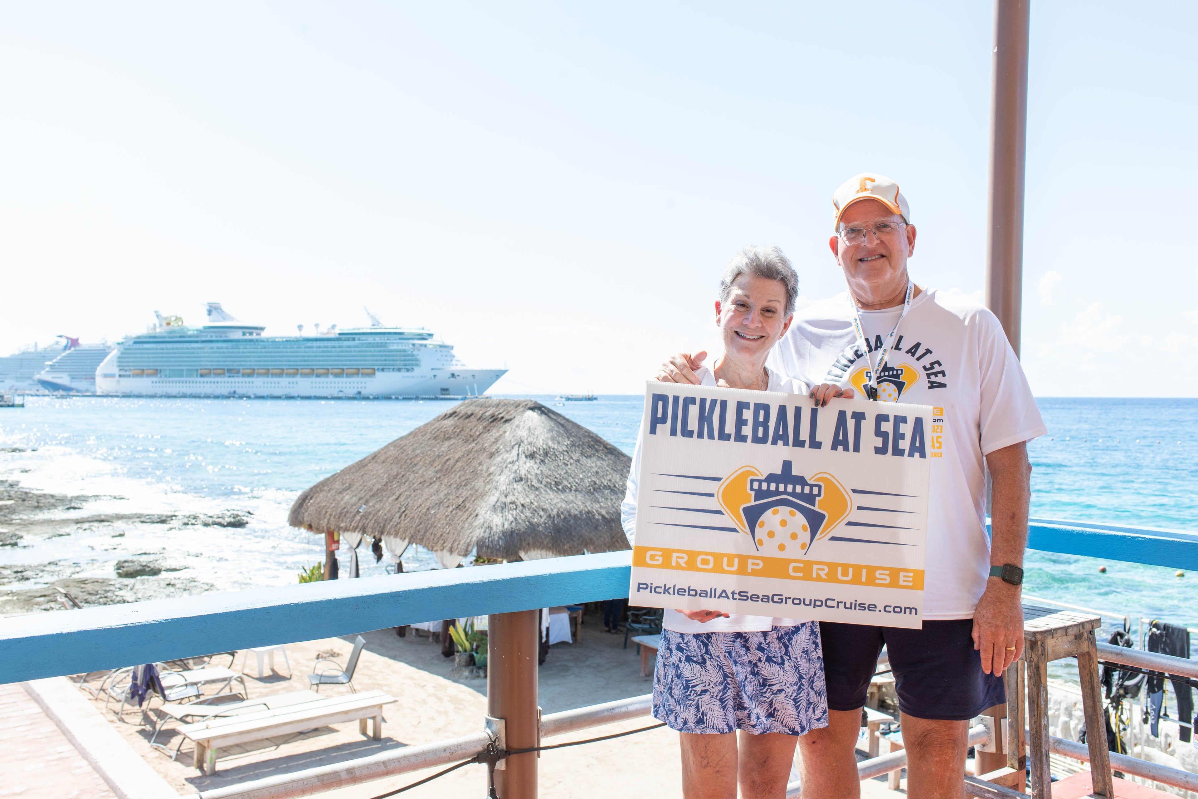 Happy couple enjoying their Pickleball vacation, holding a Pickleball At Sea Group Cruise sign on a beautiful Caribbean beach with a Royal Caribbean cruise ship in the background.